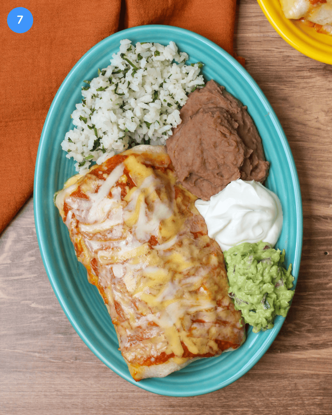 A wet ranchero burrito on a blue plate served with rice, beans, sour cream, and guacamole.