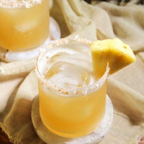 A pineapple coconut water mocktail served over ice in a rocks glass with a toasted coconut rim and a pineapple wedge garnish.
