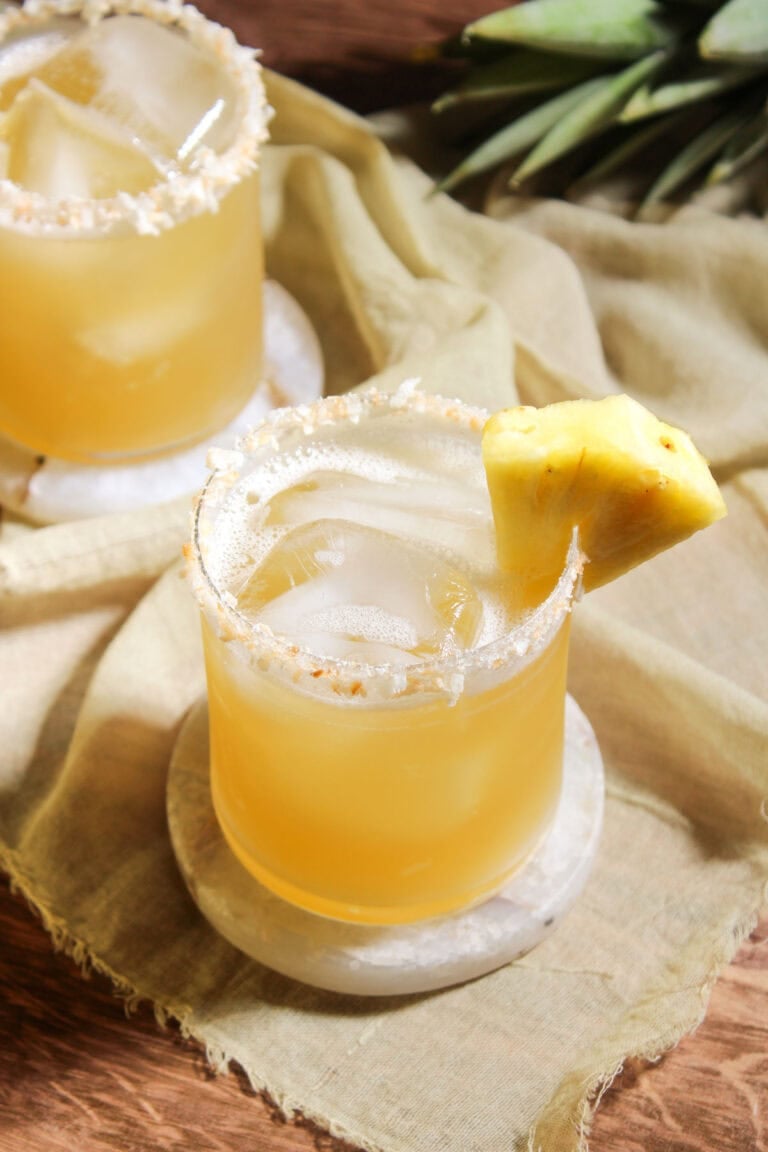A pineapple coconut water mocktail served over ice in a rocks glass with a toasted coconut rim and a pineapple wedge garnish.