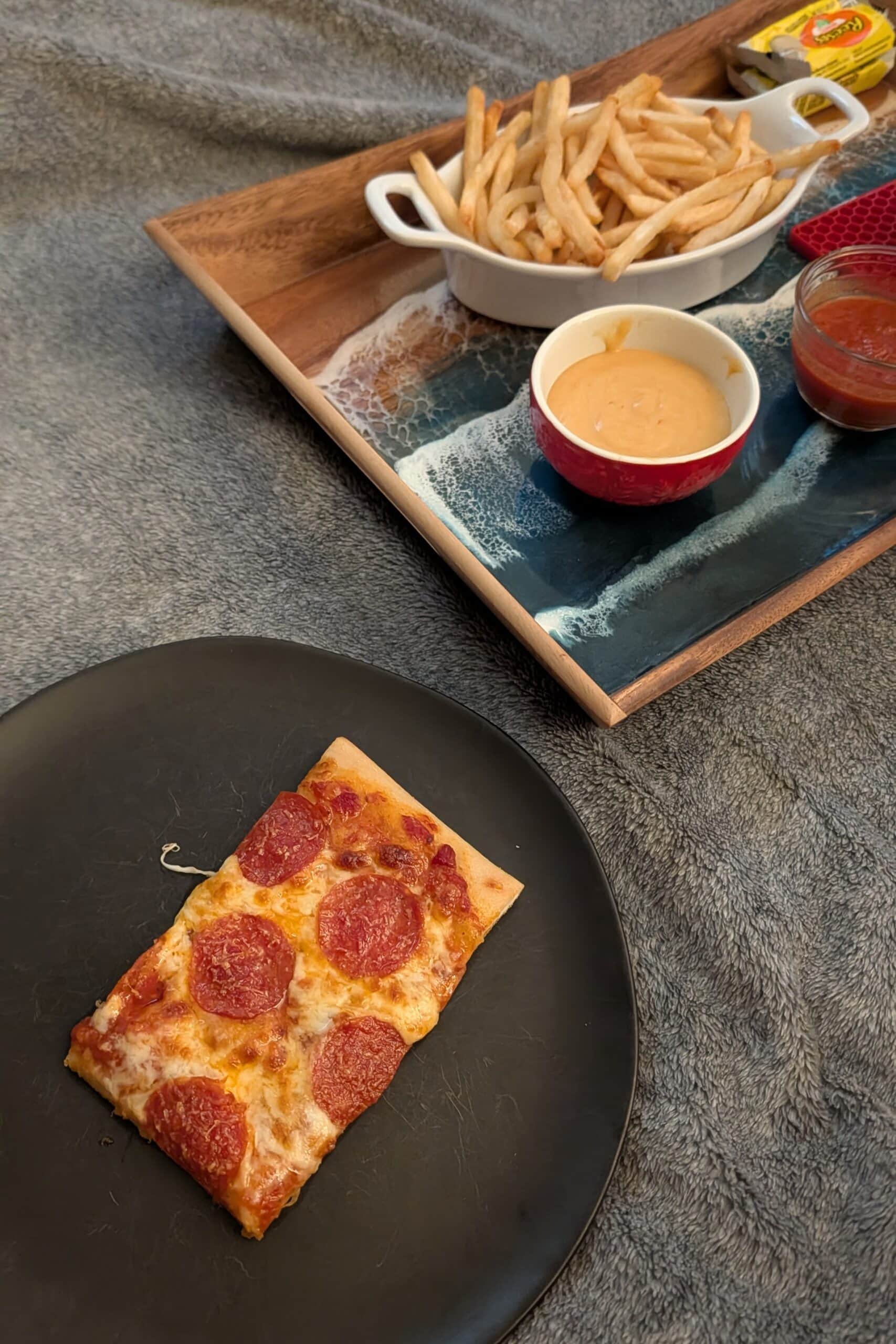 A rectangular slice of pepperoni pizza on a black plate with a tray of fries and dips.