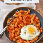 A blue stoneware bowl of freshly cooked shakshuka pasta served with an egg, with a title overlaid.