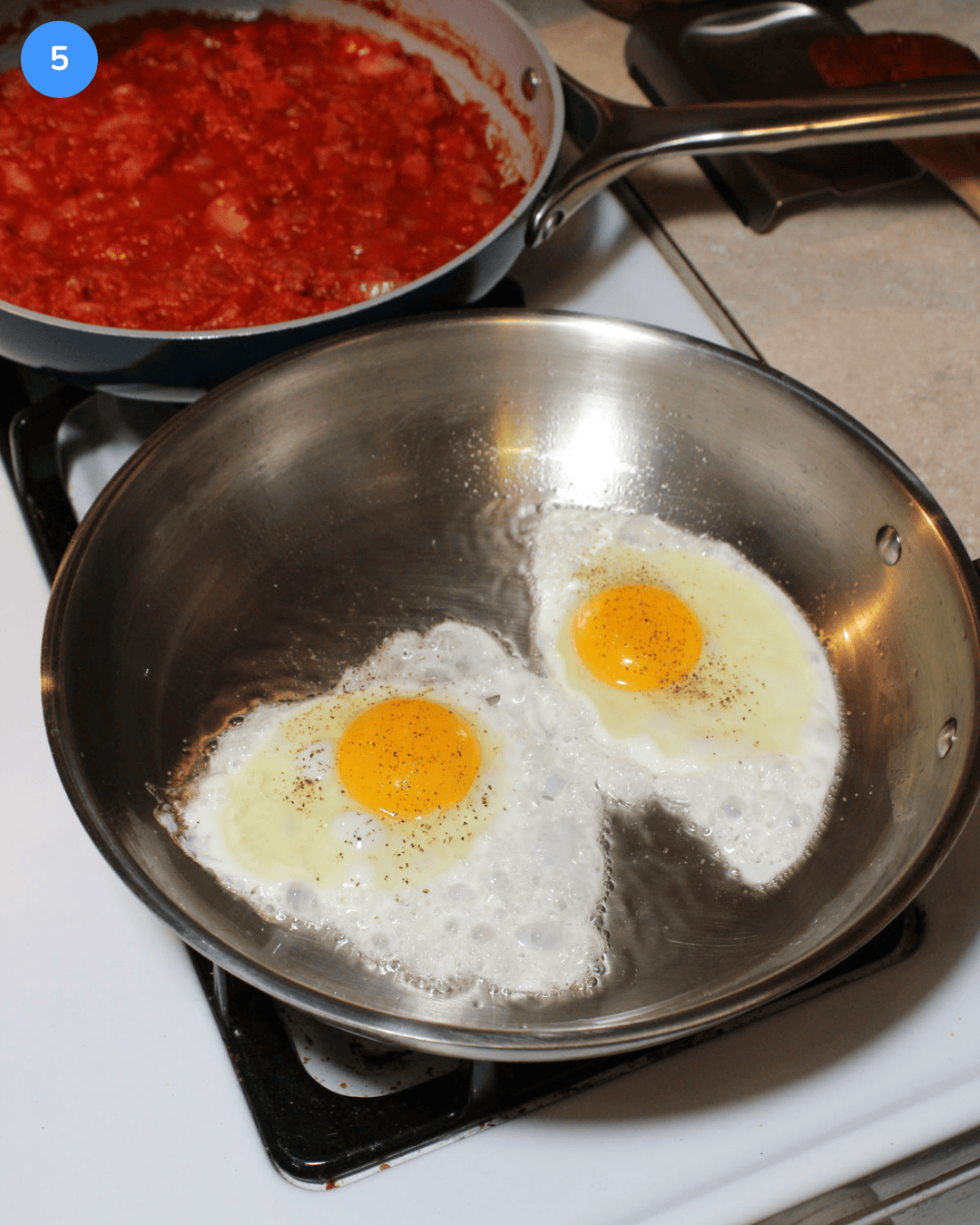 Two sunny side up eggs frying in a stainless steel pan on the stove.