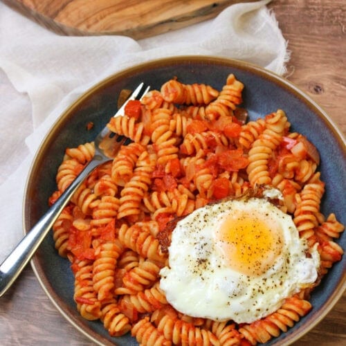 A blue stoneware bowl of shakshuka pasta served with an egg on top and freshly baked bread on the side.