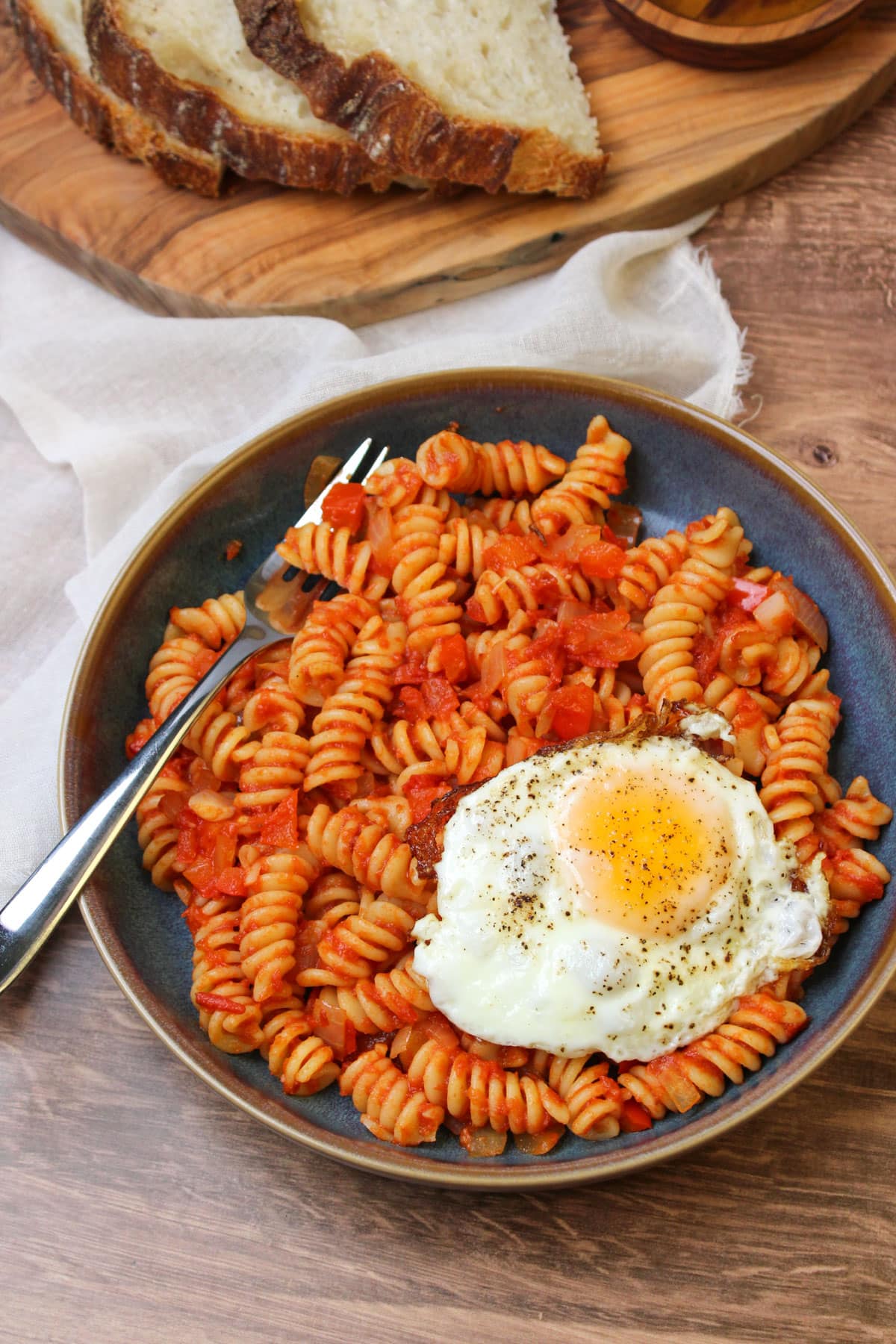 A blue stoneware bowl of shakshuka pasta served with an egg on top and freshly baked bread on the side.