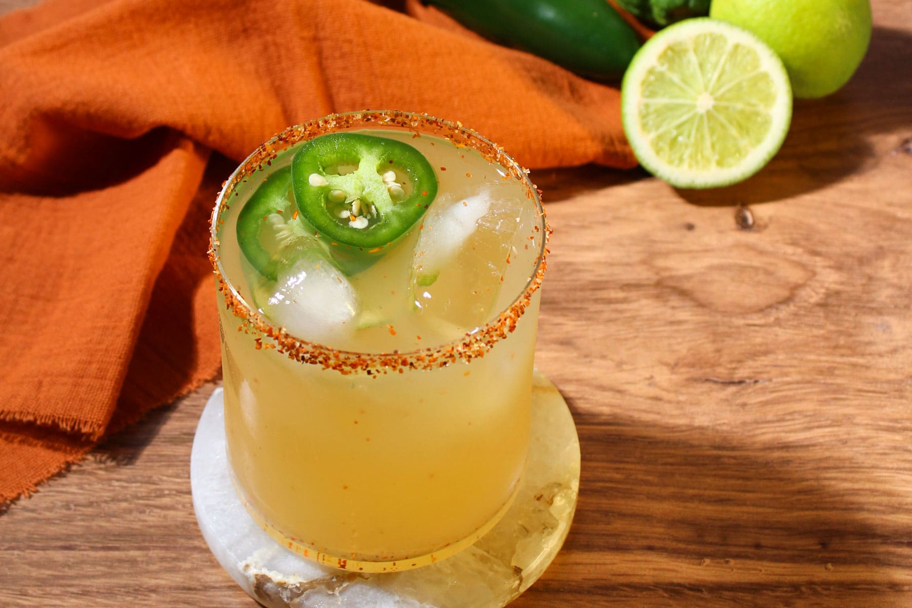 A freshly shaken mezcal margarita with a spicy rim of Tajin and a jalapeno garnish in a rocks glass on a marble coaster.