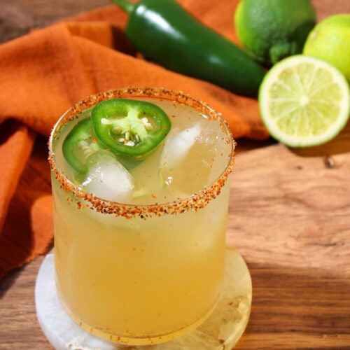 A spicy mezcal margarita in a rocks glass with a Tajin rim and jalapeno wheel garnish, on a marble coaster.