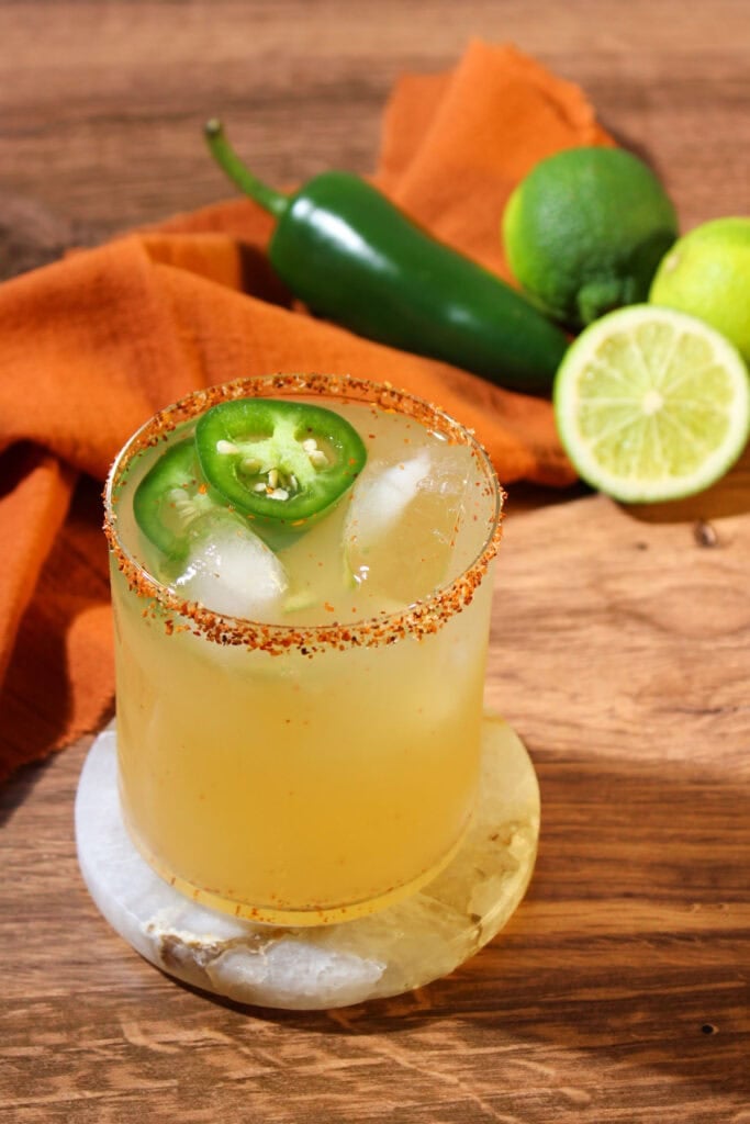 A spicy mezcal margarita in a rocks glass with a Tajin rim and jalapeno wheel garnish, on a marble coaster.