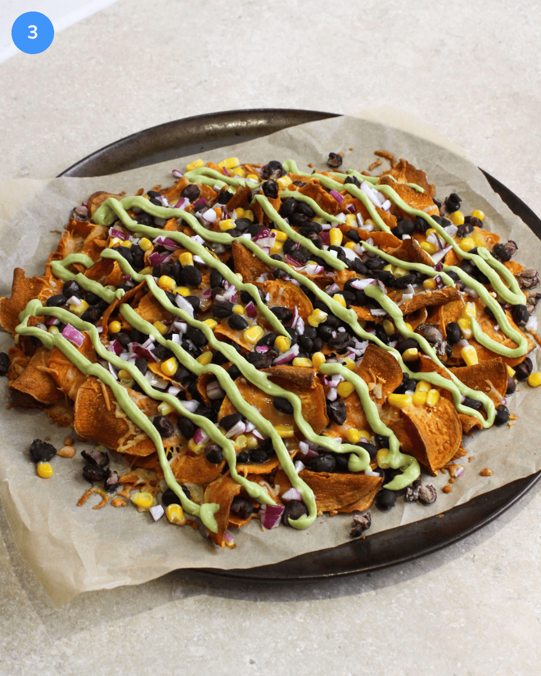 A round parchment lined pan with freshly baked sweet potato nachos, topped with gooey cheese, beans, corn, red onion, and avocado lime crema drizzle.