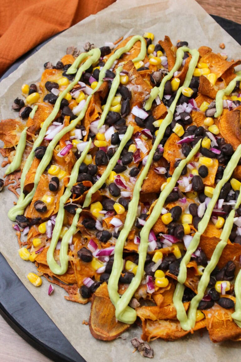 A black wooden serving platter lined with parchment paper, serving sweet potato nachos topped with melted cheese, black beans, corn, red onion, and avocado crema.