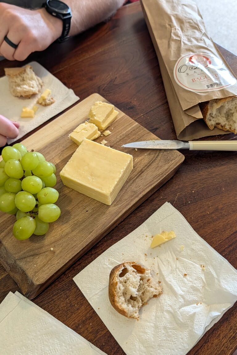 A wood cutting board with a block of cheddar cheese and green grapes, and a freshly baked baguette.