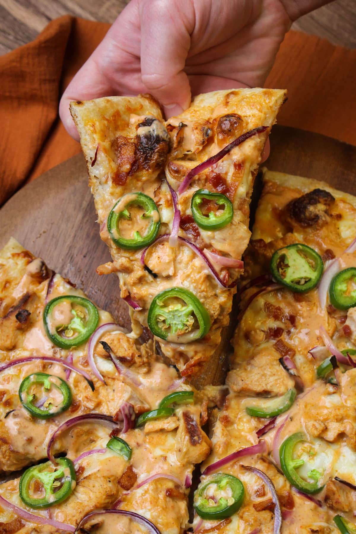 A hand picking up a slice of freshly made spicy chicken bacon ranch pizza.