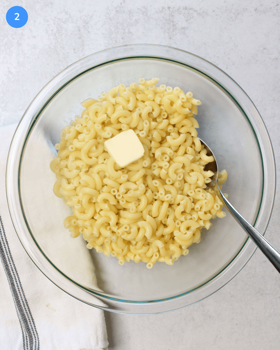 A large glass mixing bowl with freshly boiled elbow macaroni and a pad of butter in it.