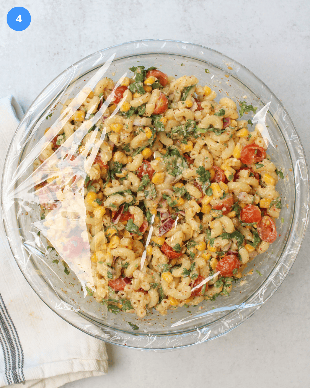 A large glass mixing bowl of elote pasta salad covered with plastic wrap.