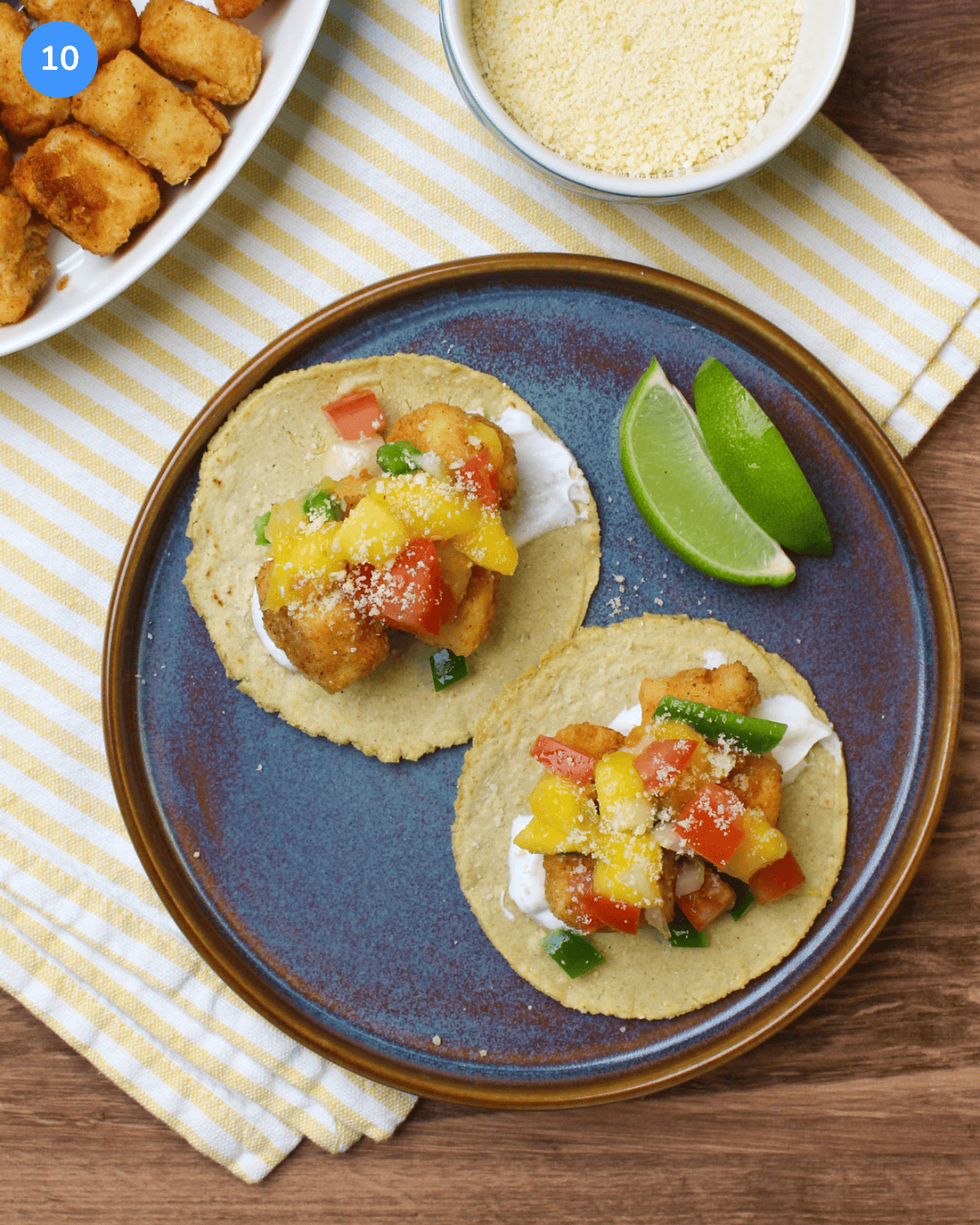 Step 10 of making fried fish tacos. Two fully assembled fried with tacos with mango salsa and lime crema on corn tortillas on a plate.