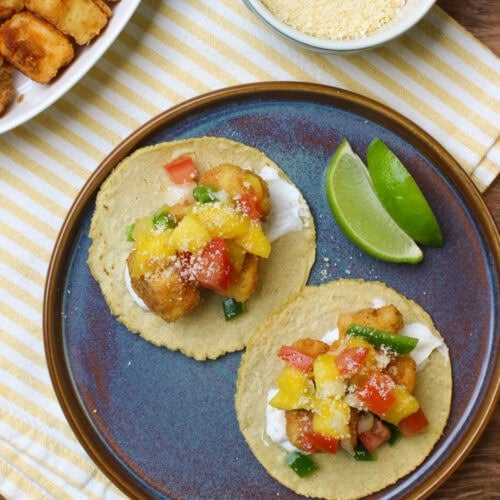 A top down image of two fried fish tacos made with corn tortillas, lime crema, fried cod, mango pico de gallo, and grated cotija and served with lime wedges on a blue stoneware plate.