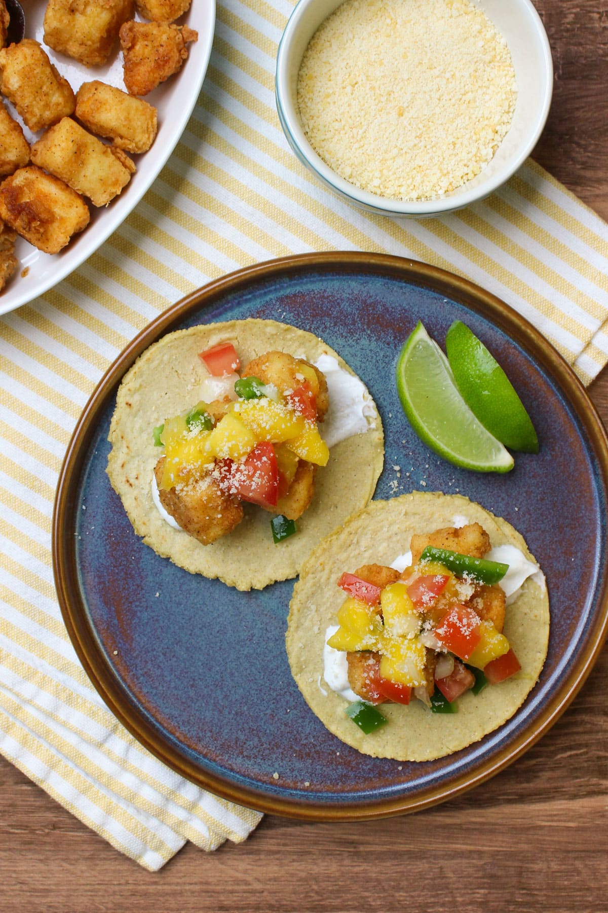 A top down image of two fried fish tacos made with corn tortillas, lime crema, fried cod, mango pico de gallo, and grated cotija and served with lime wedges on a blue stoneware plate.