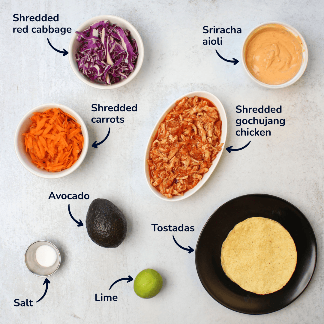 Ingredients for making gochujang chicken tostadas: shredded red cabbage, shredded carrots, lime, salt, avocado, tostadas, shredded gochujang chicken, and sriracha aioli.