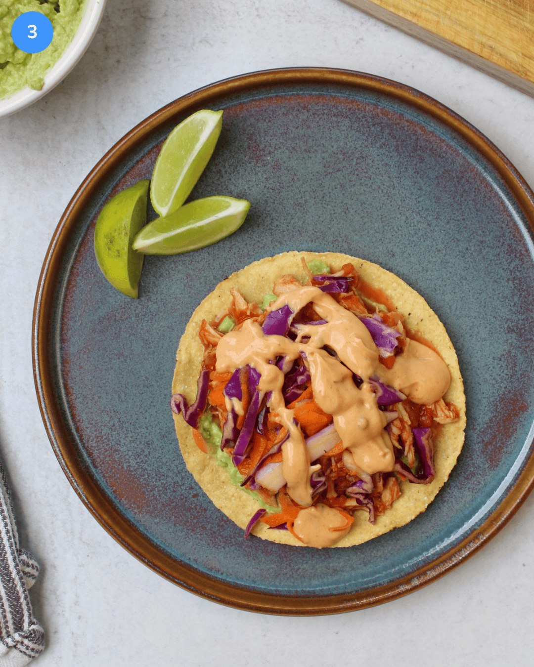 A freshly made gochujang chicken tostada on a blue ceramic plate, topped with sriracha aioli and served with lime wedges.