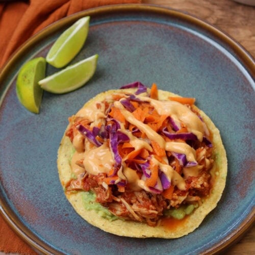 A Korean Mexican fusion gochujang chicken tostada served on a blue stoneware plate with lime wedges.
