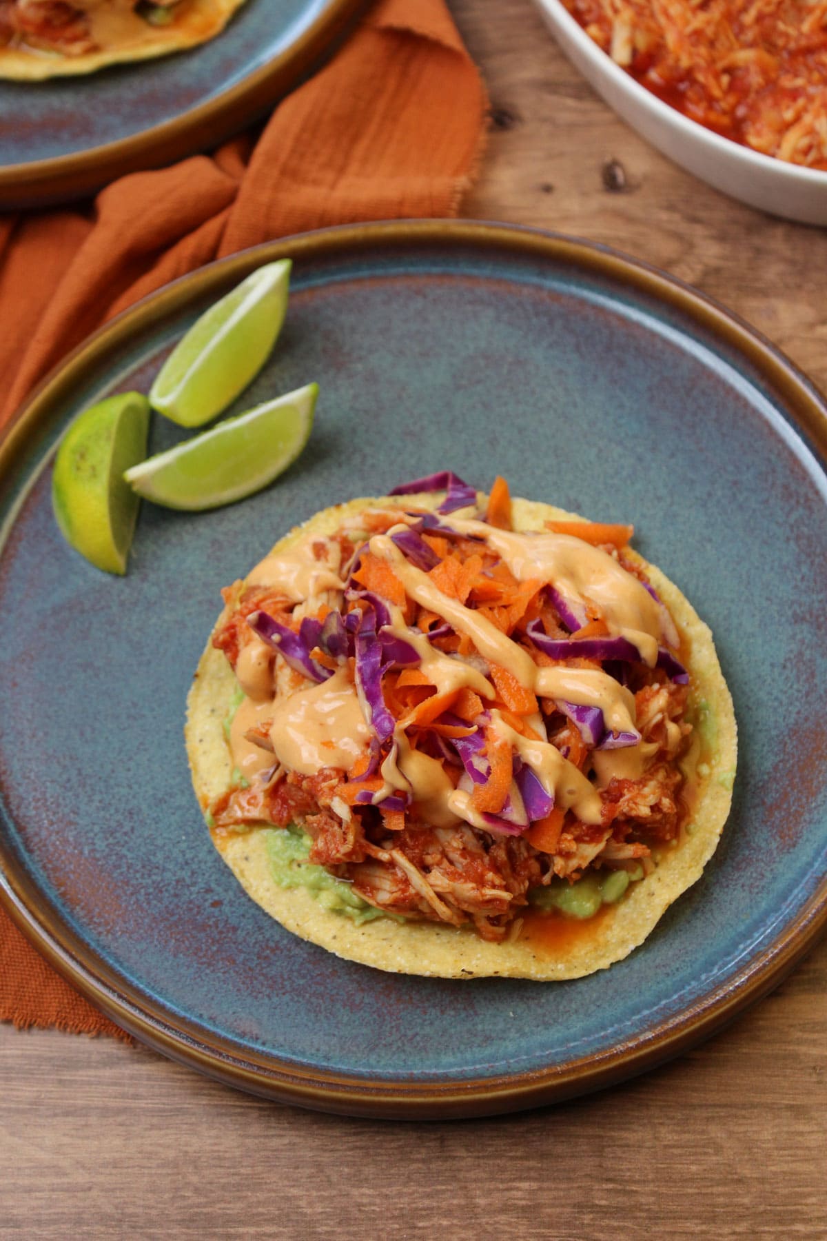 A Korean Mexican fusion gochujang chicken tostada served on a blue stoneware plate with lime wedges.