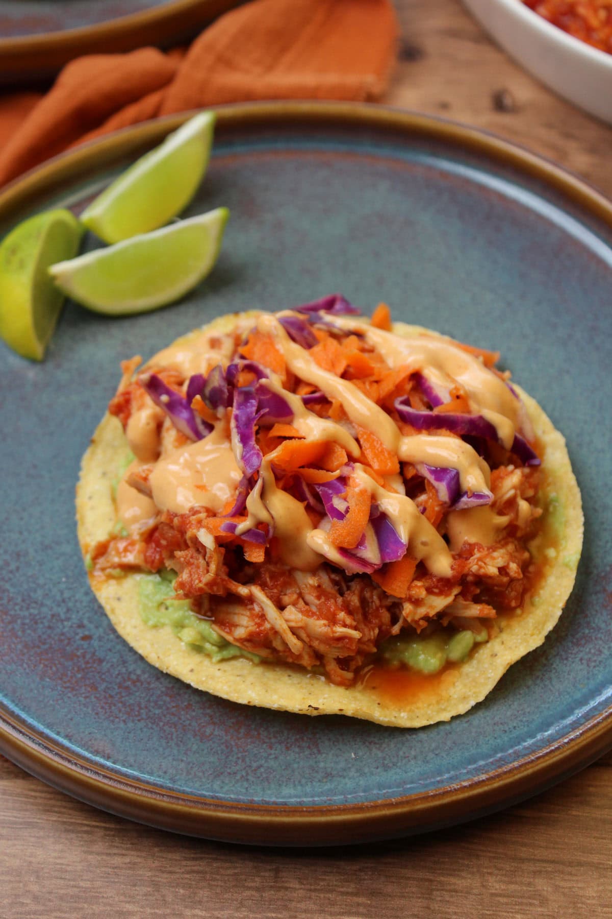 A shredded gochujang chicken tostada topped with carrot and cabbage slaw and sriracha mayo, served on a blue stone plate with lime wedges.