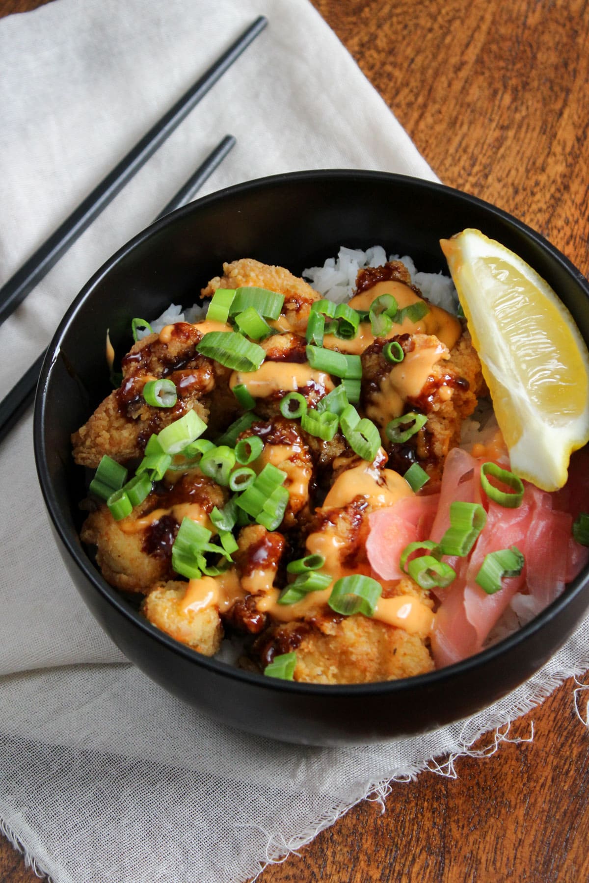 A black bowl of freshly made Japanese crispy chicken and rice with sesame soy glaze, sriracha aioli, pickled ginger, scallions, and a lemon wedge.