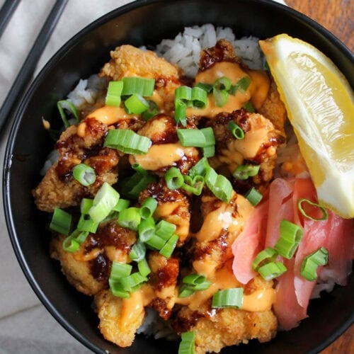 A freshly prepared Japanese crispy chicken rice bowl with soy glaze, sriracha aioli, scallions, pickled ginger, and a lemon wedge.