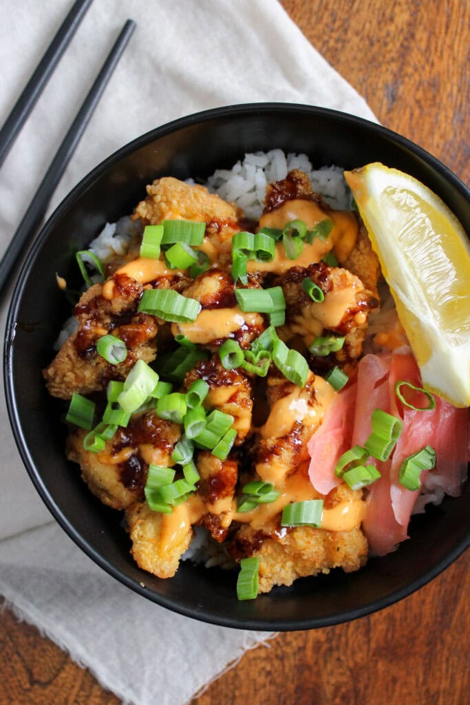 A freshly prepared Japanese crispy chicken rice bowl with soy glaze, sriracha aioli, scallions, pickled ginger, and a lemon wedge.