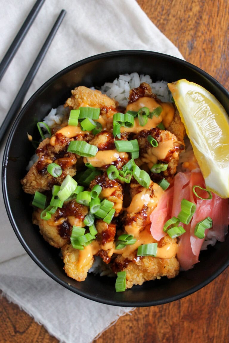 A freshly prepared Japanese crispy chicken rice bowl with soy glaze, sriracha aioli, scallions, pickled ginger, and a lemon wedge.