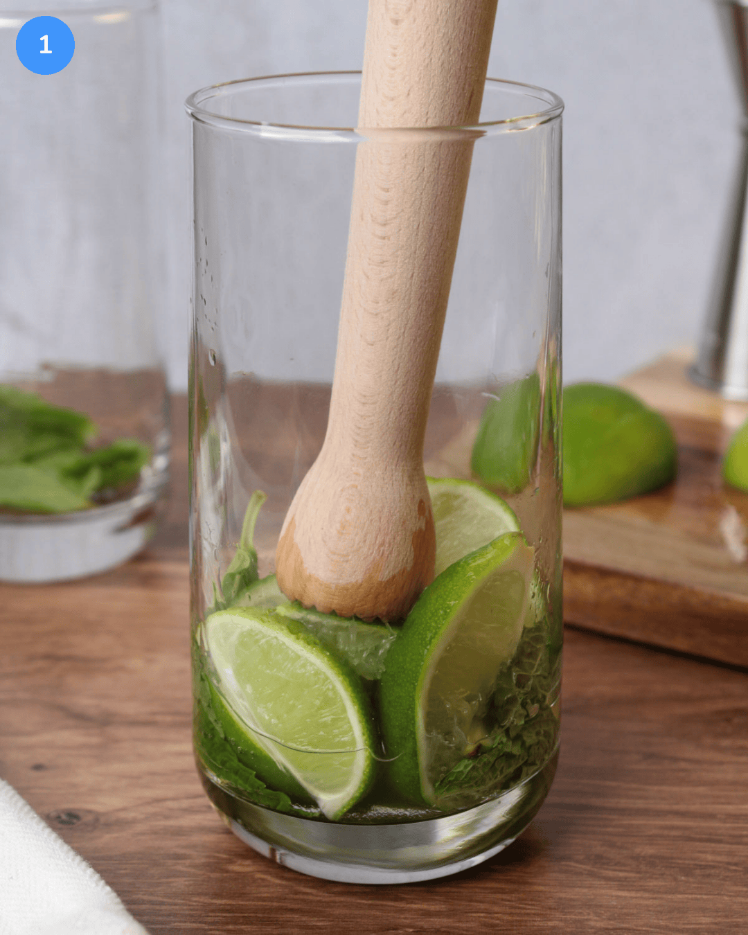 Fresh mint, lime wedges, and simple syrup being muddled in a Collins glass.