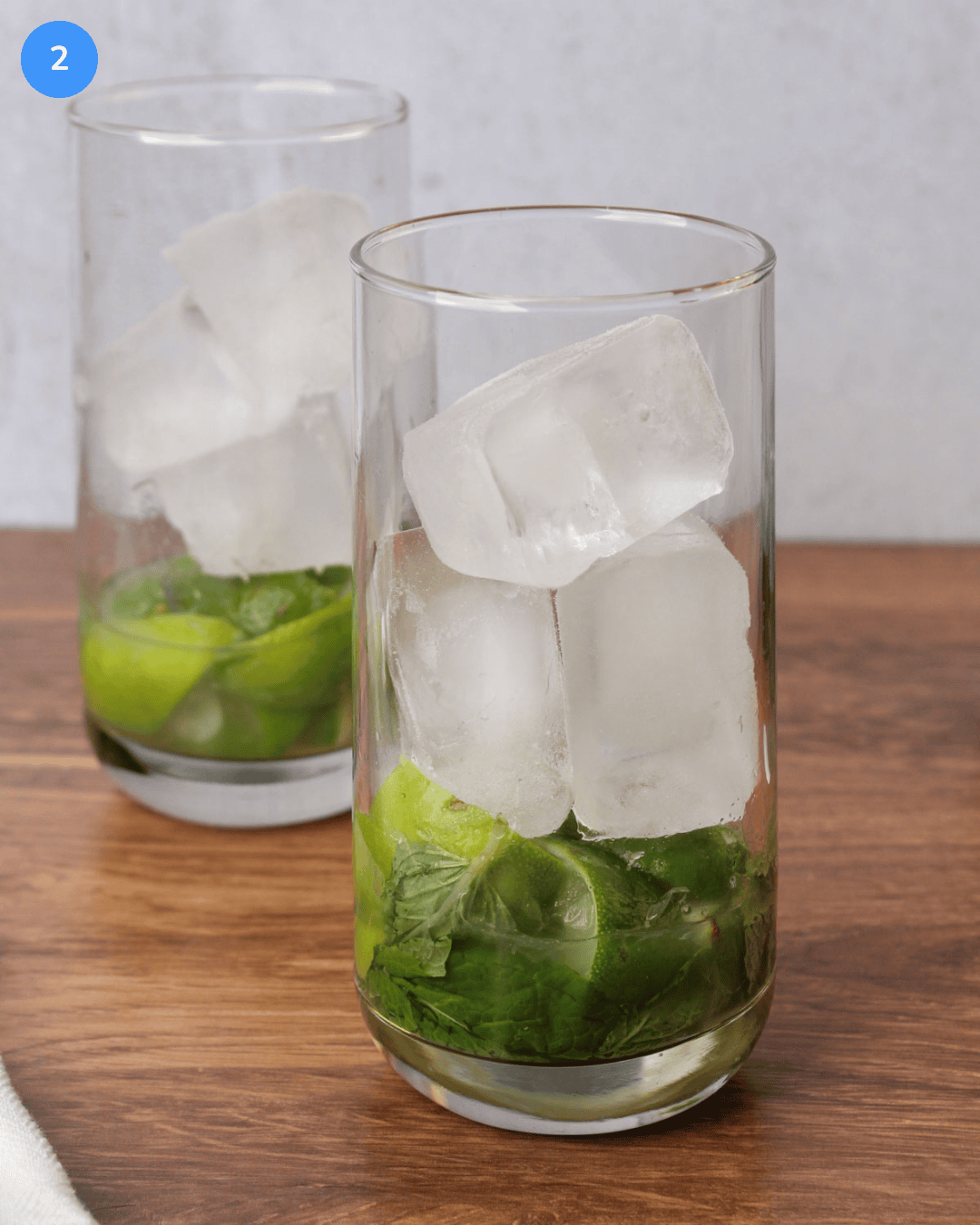 A collins glass filled with muddled mint, lime wedges, simple syrup, and large ice cubes.