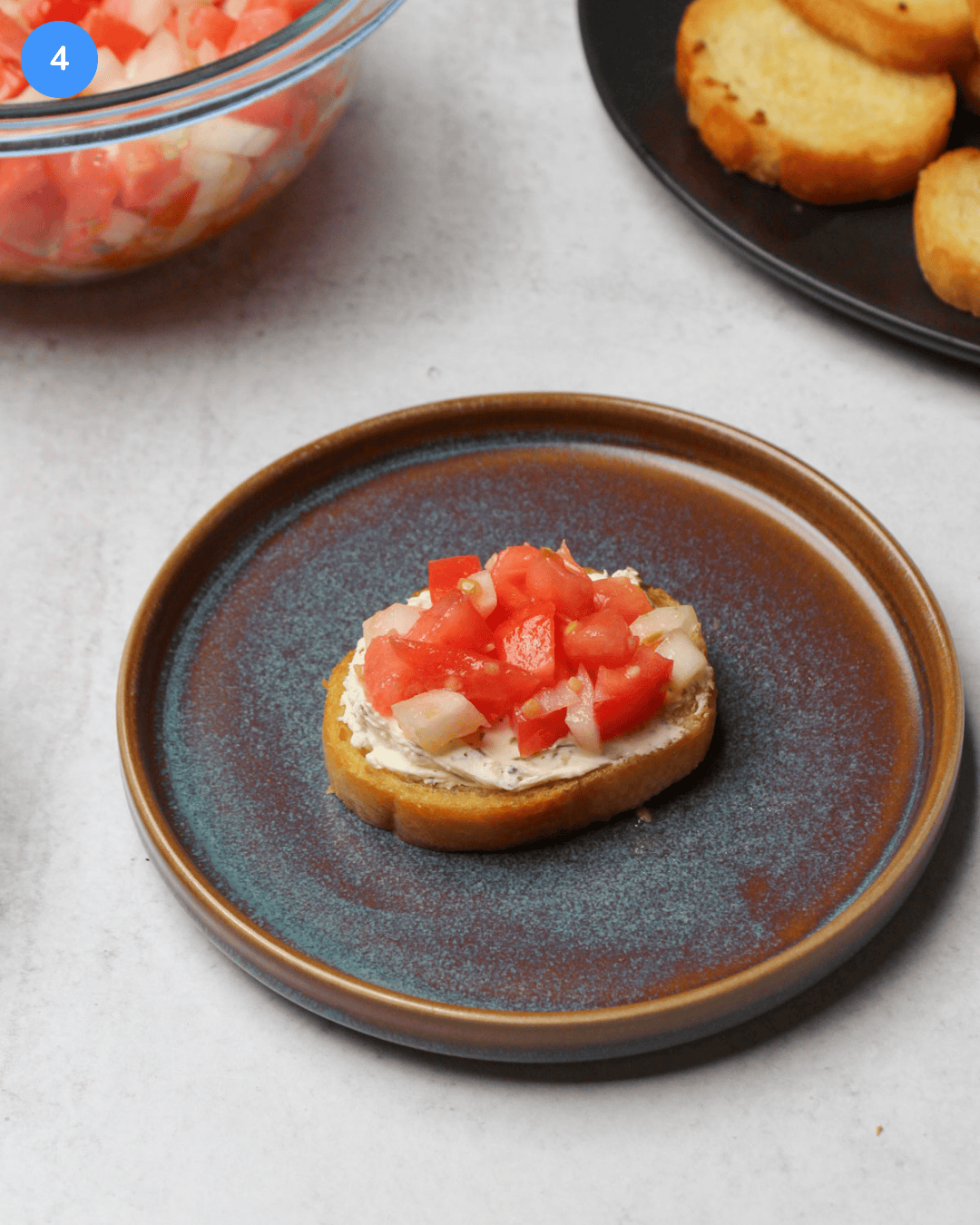A small crostini topped with garlic herb spreadable cheese and tomato bruschetta served on a small blue stone plate.