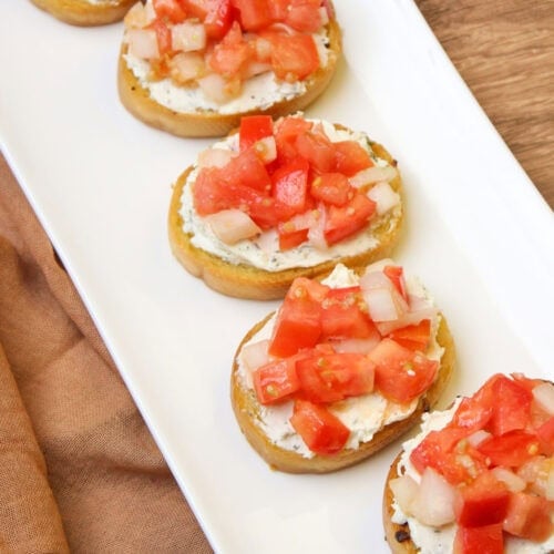 A long white serving platter of tomato bruschetta crostini with alouette garlic and herb cheese spread.