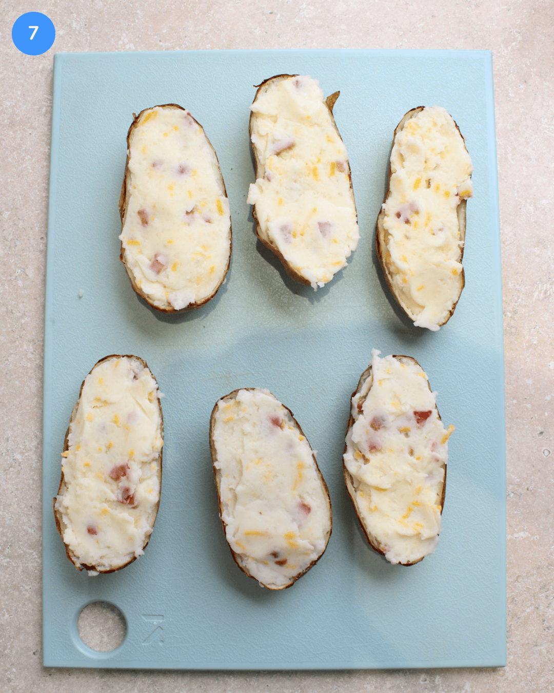 Step 7: 6 air fried potato halves that have been filled with a potato, cheese, bacon, butter, and heavy cream mixture on a cutting board.