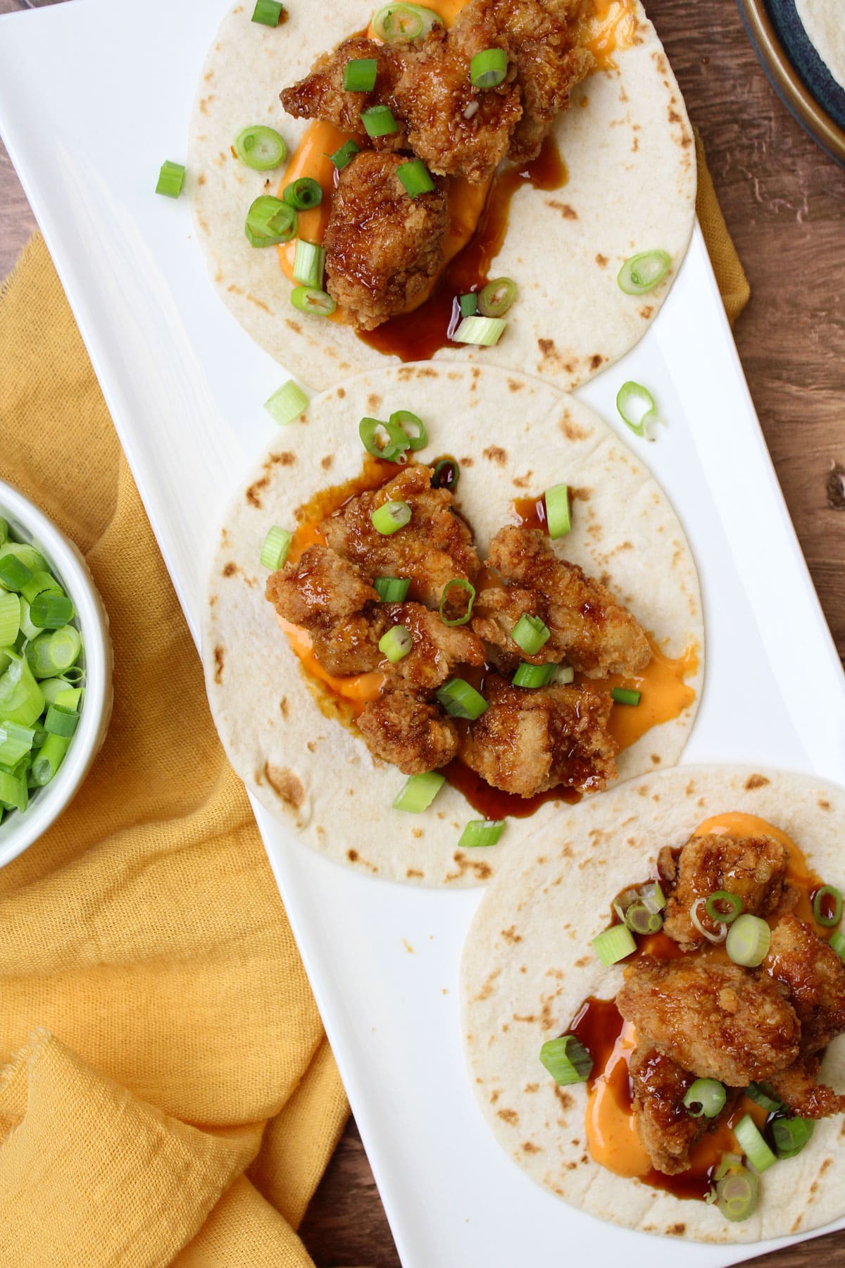 A long white plate of fried chicken tacos on flour tortillas, freshly garnished with scallions.
