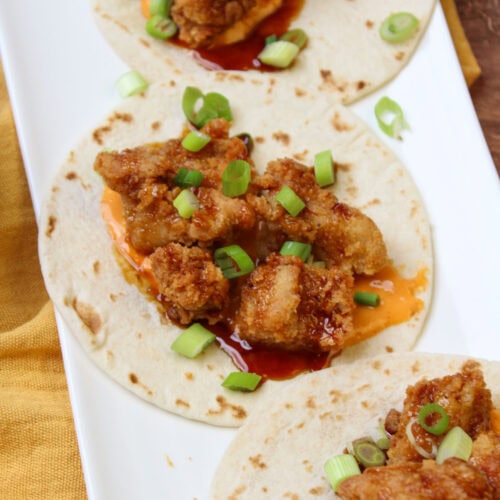 A white plate of freshly made fried chicken tacos, topped with asian sesame glaze, sriracha aioli, and scallions.