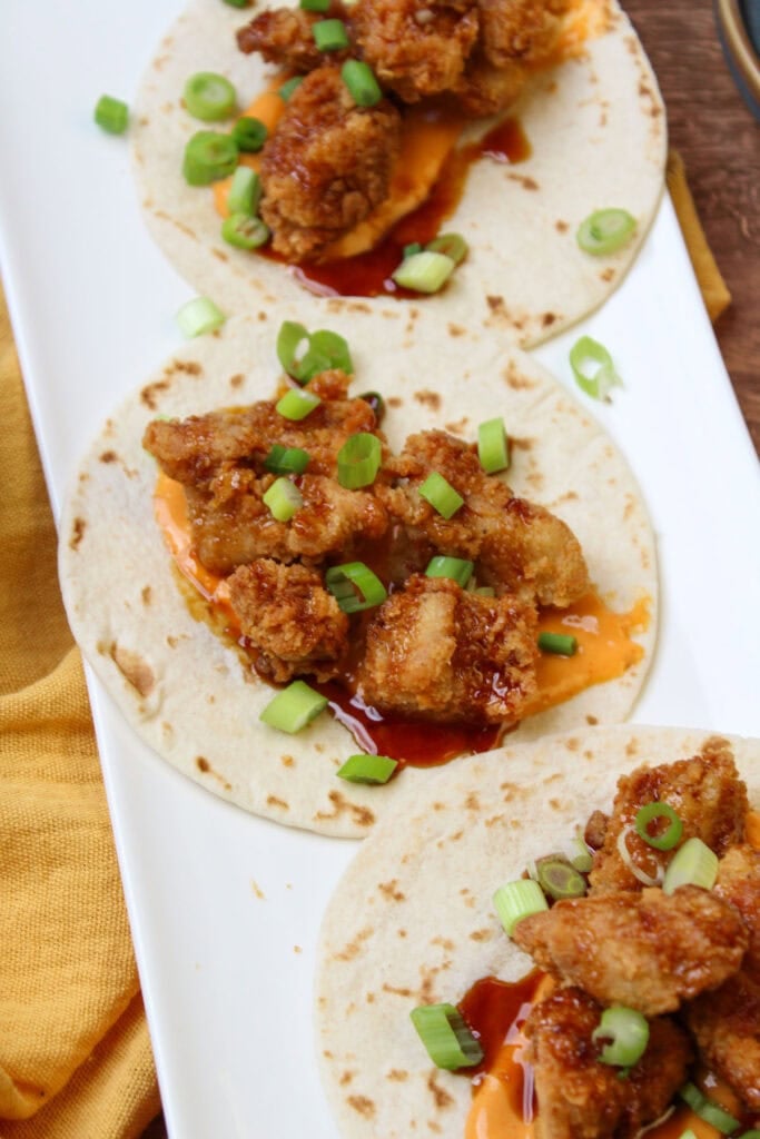 A white plate of freshly made fried chicken tacos, topped with asian sesame glaze, sriracha aioli, and scallions.
