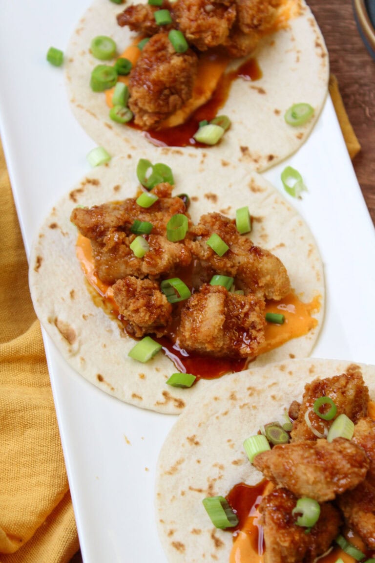 A white plate of freshly made fried chicken tacos, topped with asian sesame glaze, sriracha aioli, and scallions.