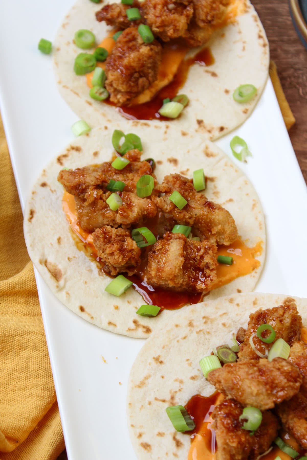 A white plate of freshly made fried chicken tacos, topped with asian sesame glaze, sriracha aioli, and scallions.