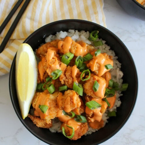 A black bowl of freshly made fried fish with rice, sriracha aioli, scallions, and a lemon wedge.