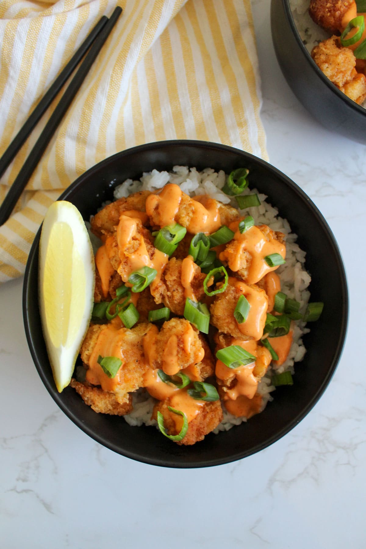 A black bowl of freshly made fried fish with rice, sriracha aioli, scallions, and a lemon wedge.