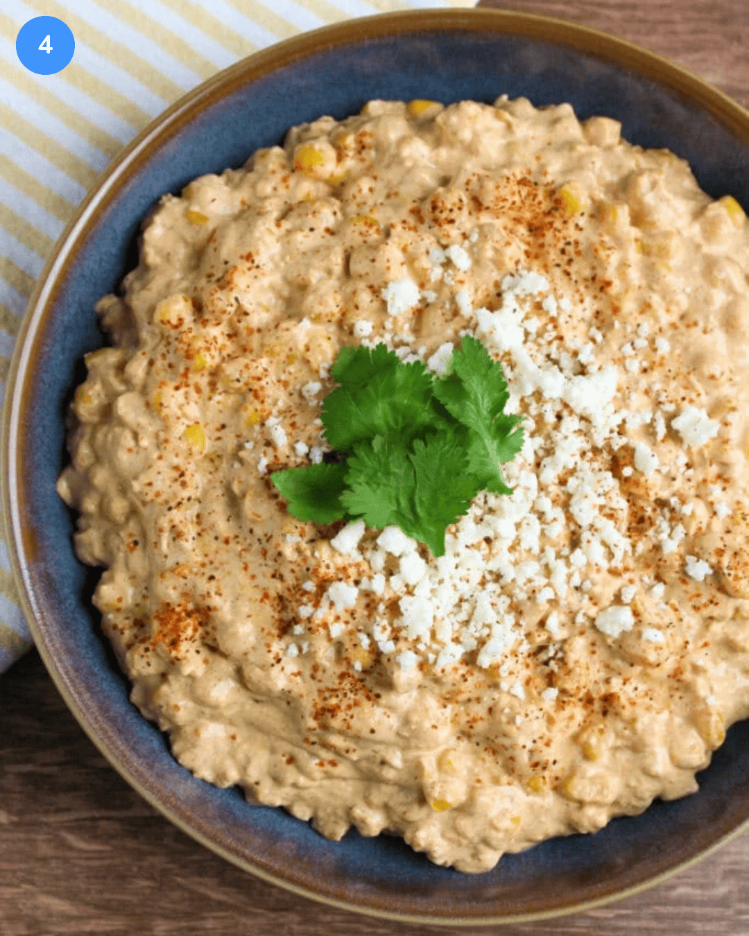 Step 4 of Mexican street corn dip: The mexican street corn dip is plated in a blue stoneware bowl and topped with Tajin, cotija, and fresh cilantro for garnish.