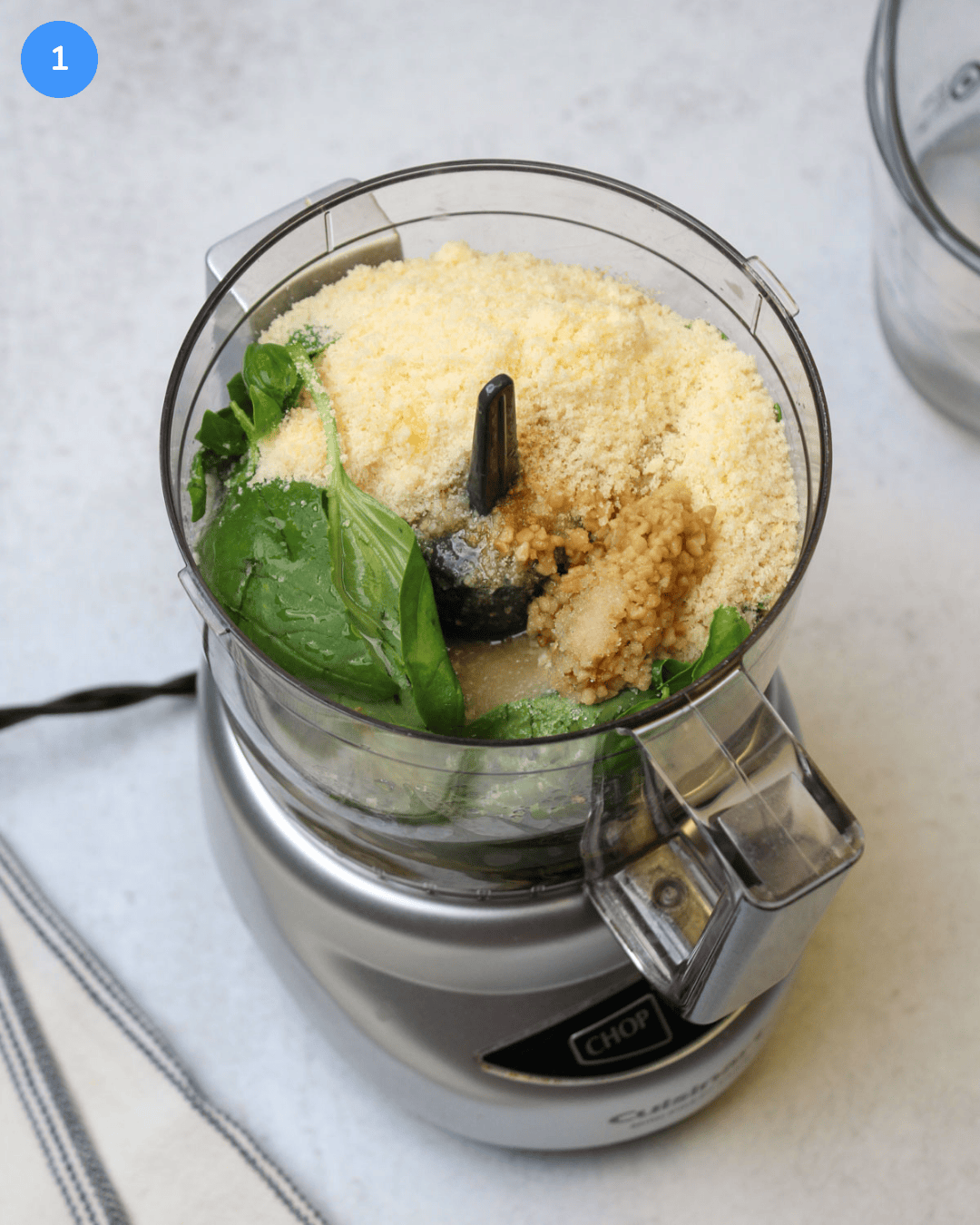 A food processor filled with fresh basil, breadcrumbs, grated parmesan, olive oil, minced garlic, and salt.