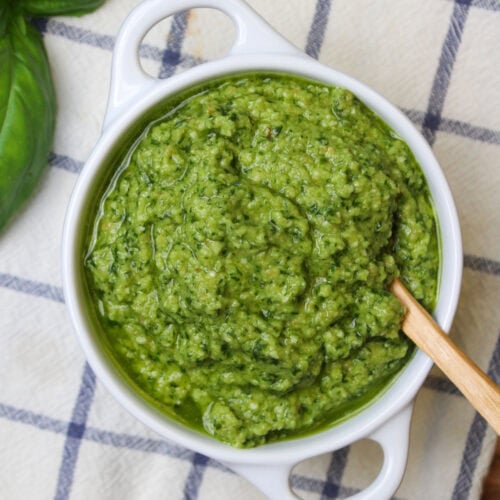 A white ceramic bowl of fresh basil pesto without nuts, with a wooden serving spoon.