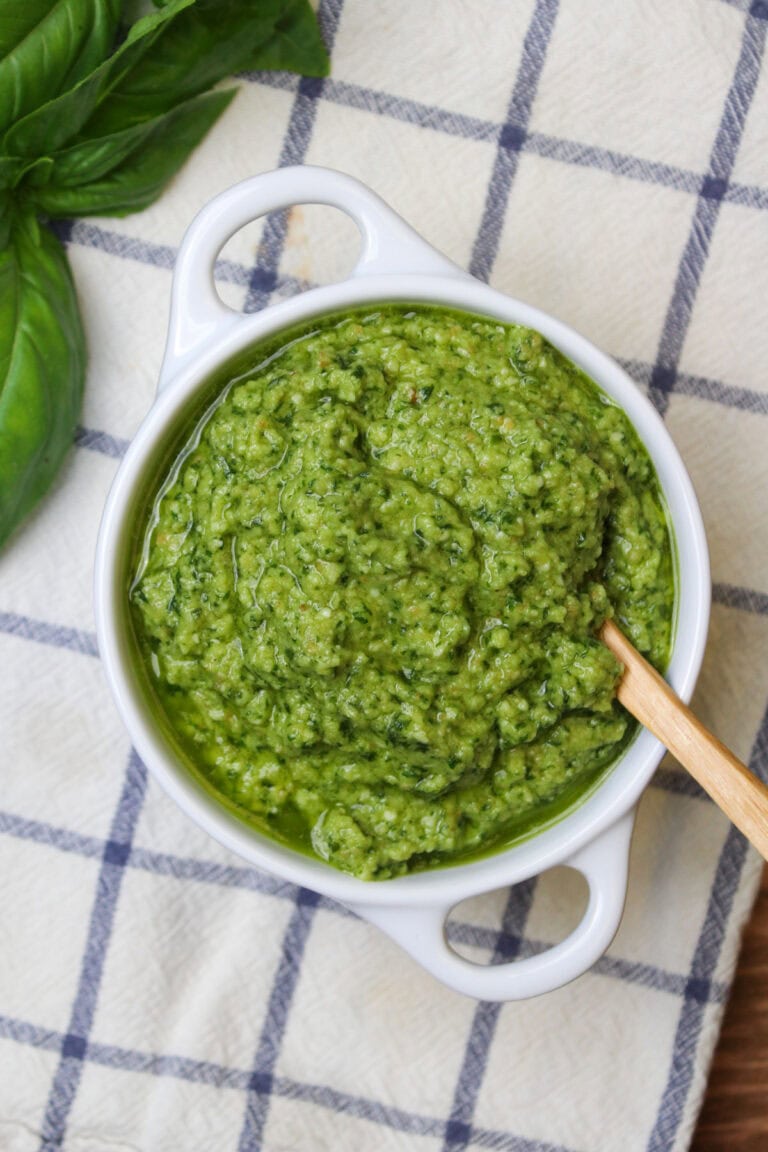 A white ceramic bowl of fresh basil pesto without nuts, with a wooden serving spoon.