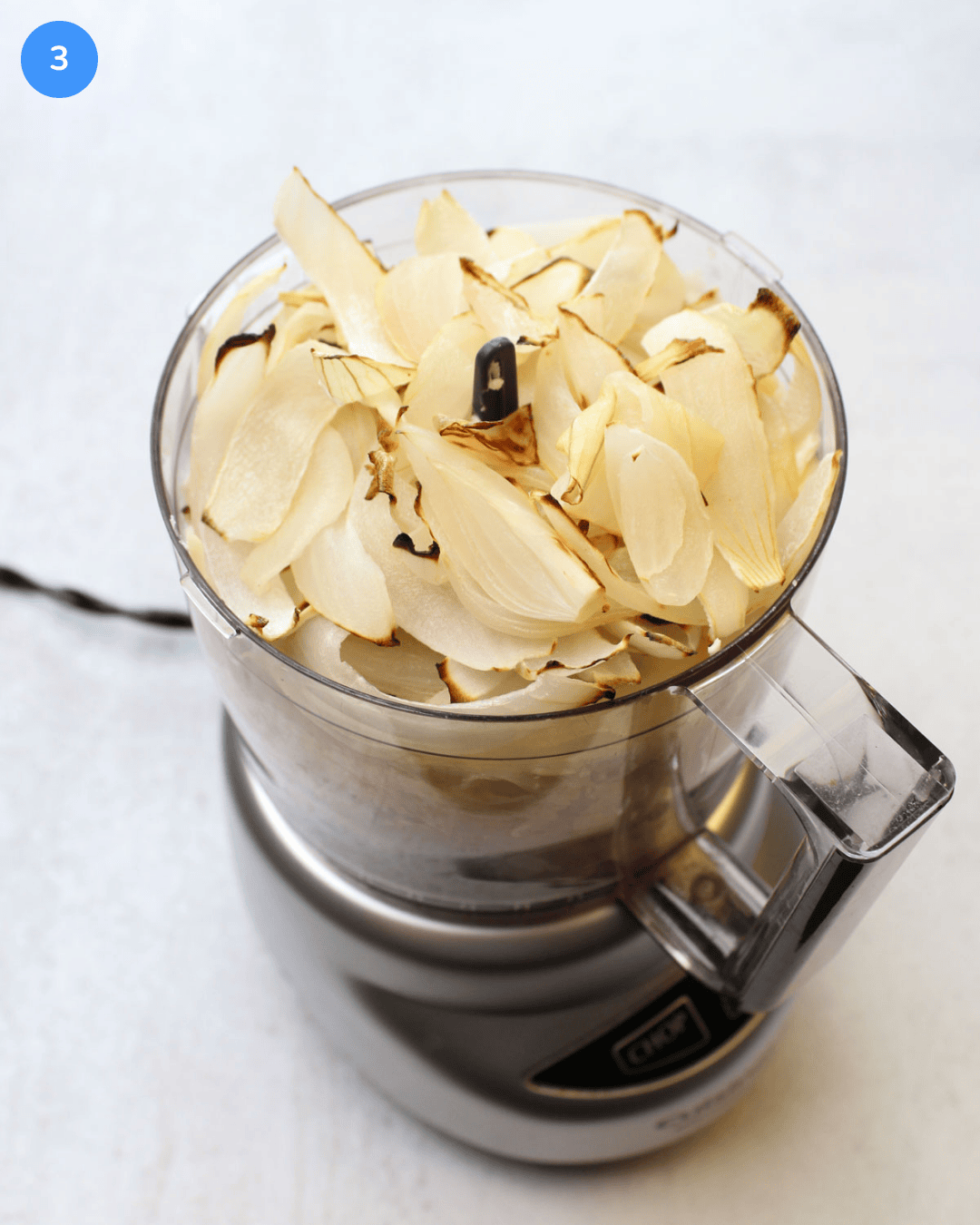 A food processor filled with cream cheese, cheese, garlic, salt, greek yogurt, and charred vidalia onion slices.