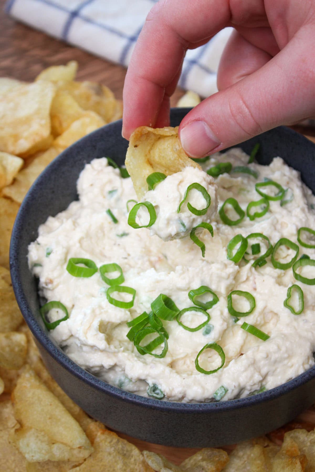 A hand dipping a kettle chip into a freshly made bowl of creamy onion dip topped with scallions.