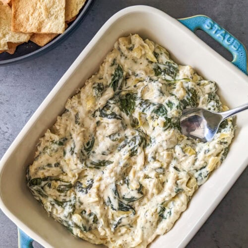 A white ceramic baking dish of spinach and artichoke dip served with pita chips.
