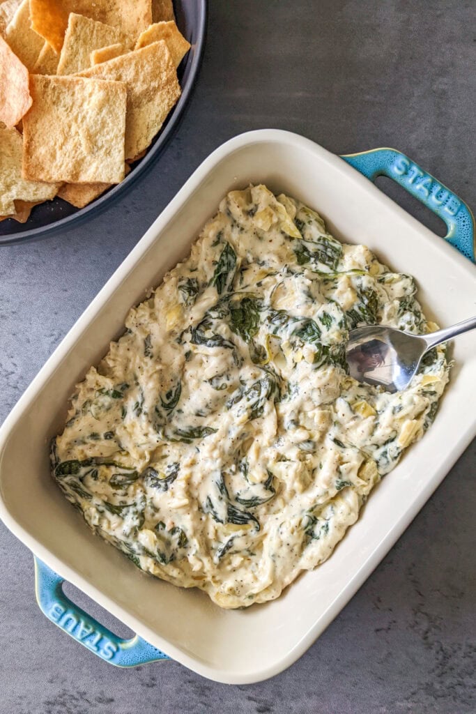 A white ceramic baking dish of spinach and artichoke dip served with pita chips.