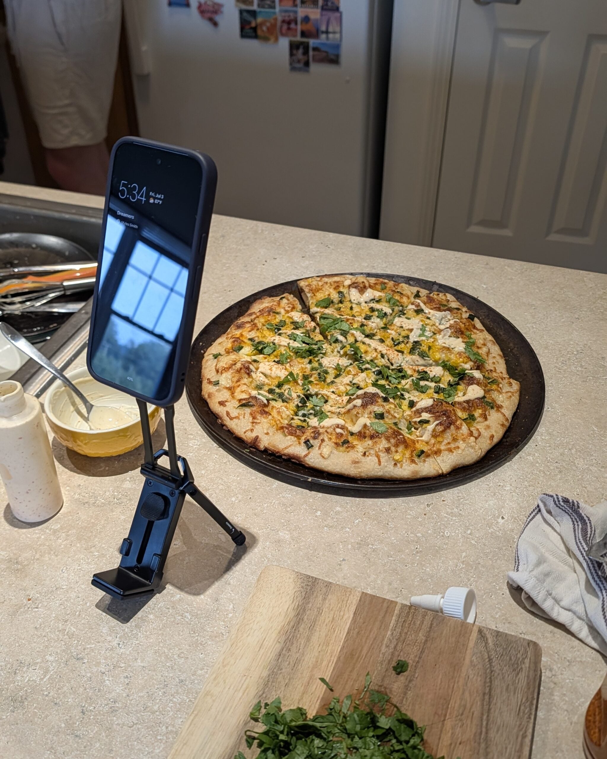 A kitchen scene with a pizza and a phone on a tripod for filming.